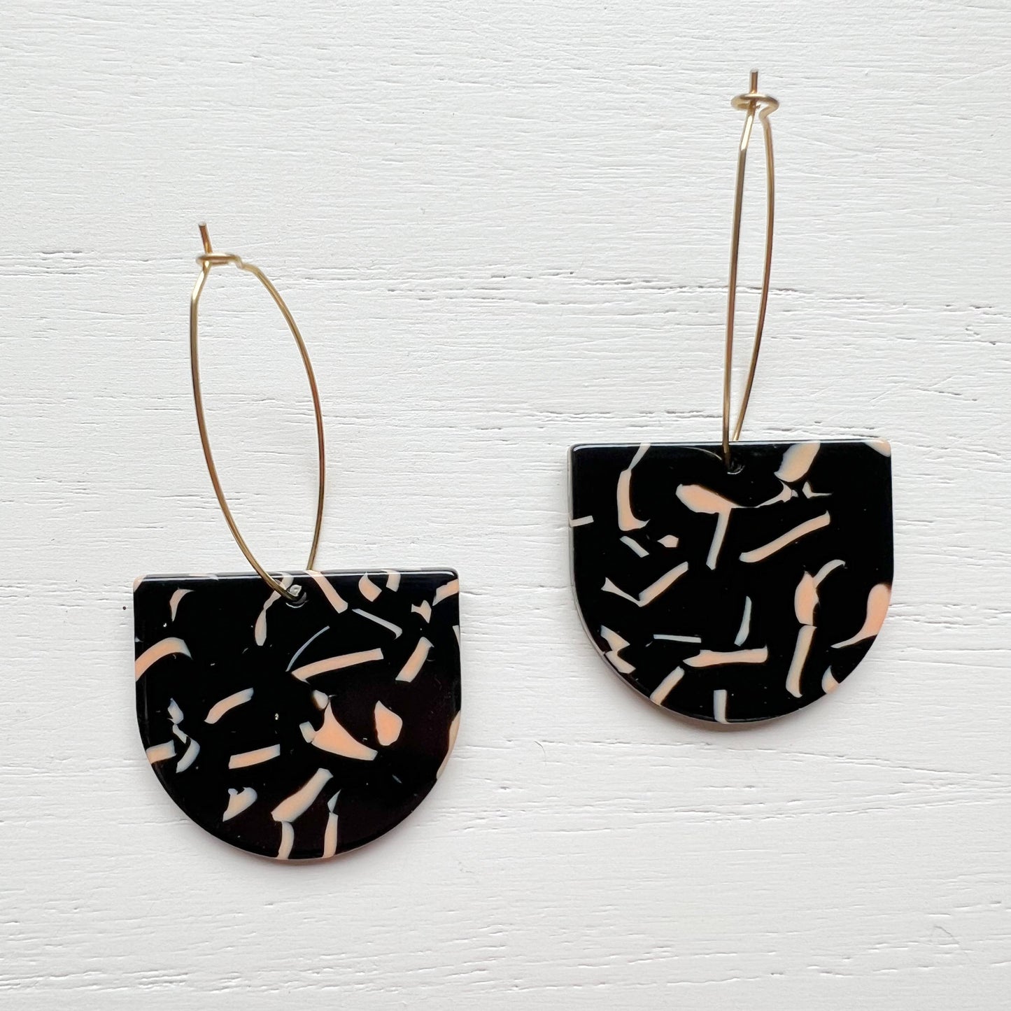 A pair of geometric drop earrings featuring black terrazzo-patterned semi-circular pendants with gold wire hoops, displayed against a white painted wood background.