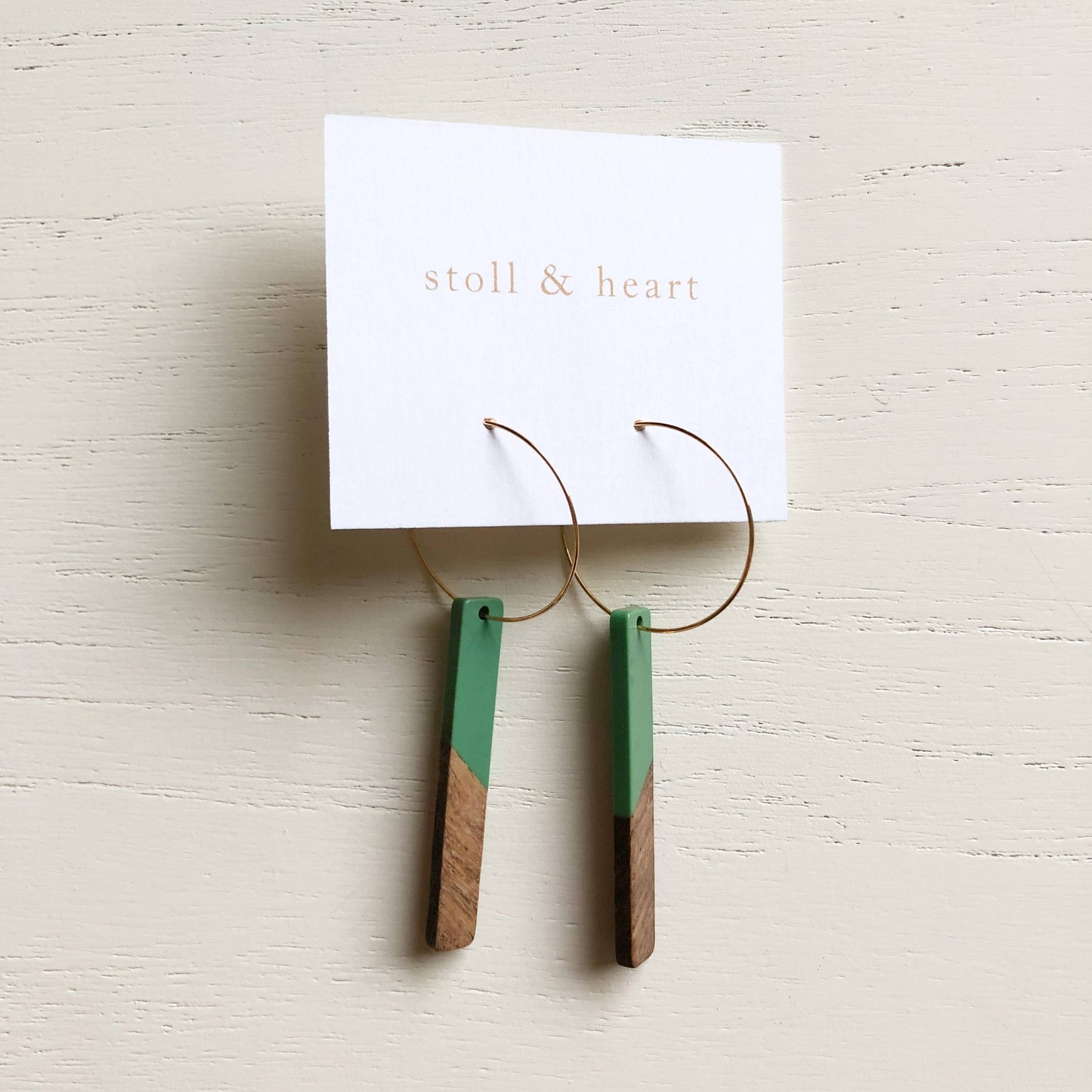 Pair of earrings with green and brown design on a white card with 'stoll & heart' branding, on a light wooden surface.