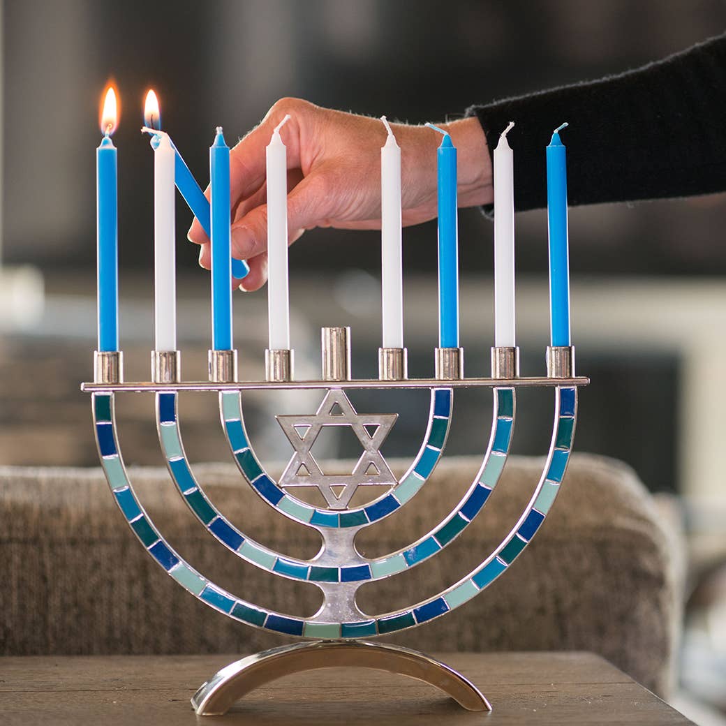 Mosaic menorah with lit candles being adjusted by a hand on a wooden surface.