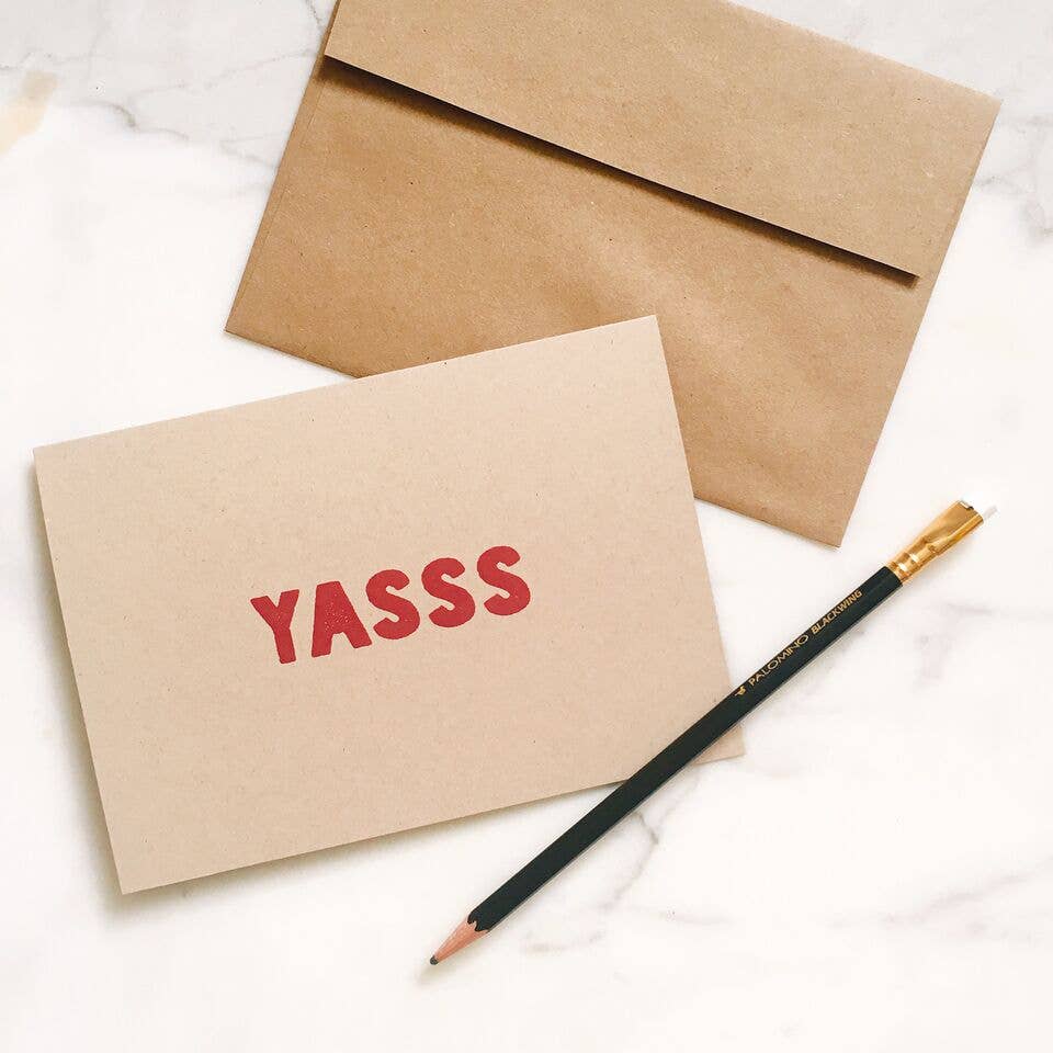 Kraft paper card with 'YASSS' text, brown envelope, and pencil on a white marble surface