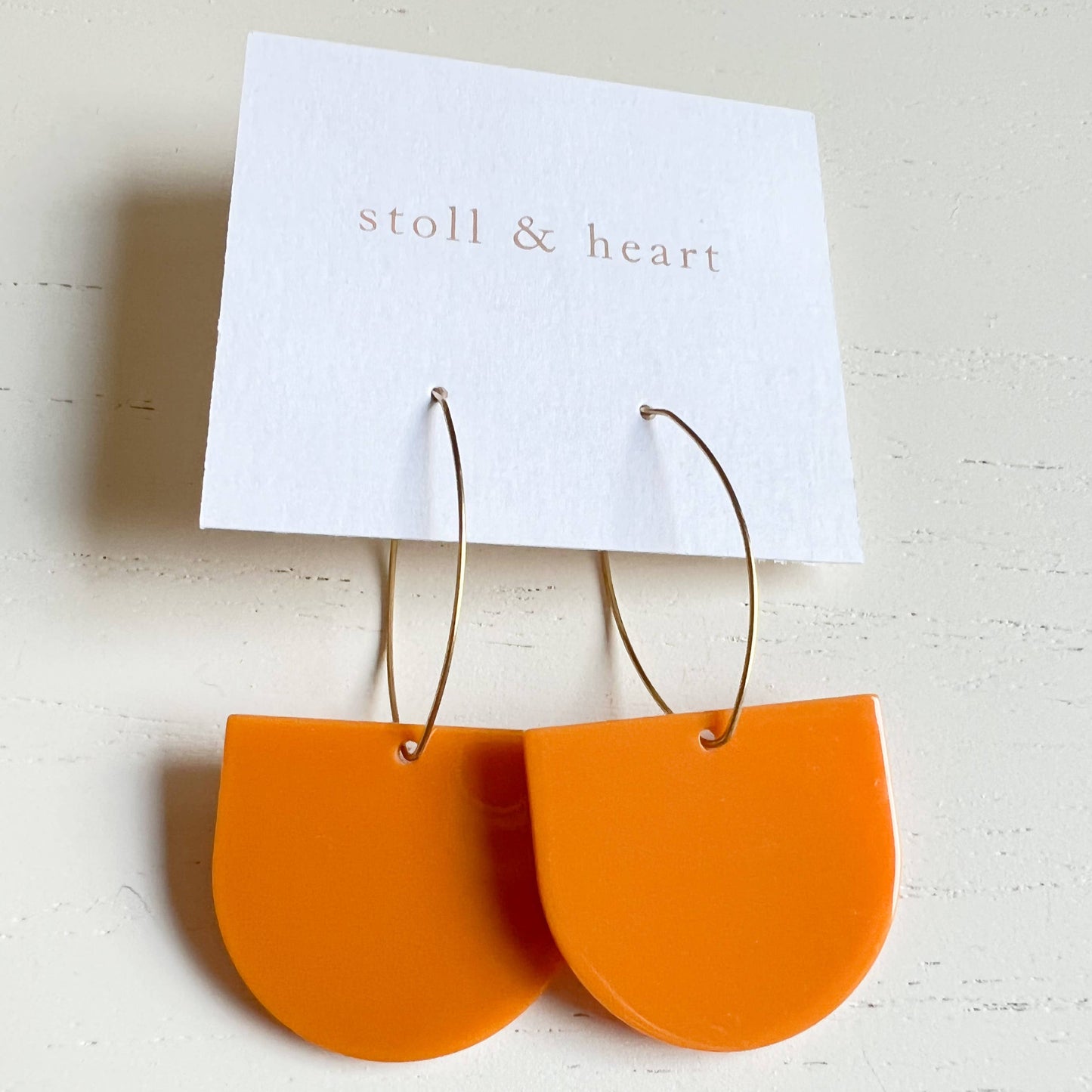 Orange half-circle earrings on a 'stoll & heart' branded card against a light background