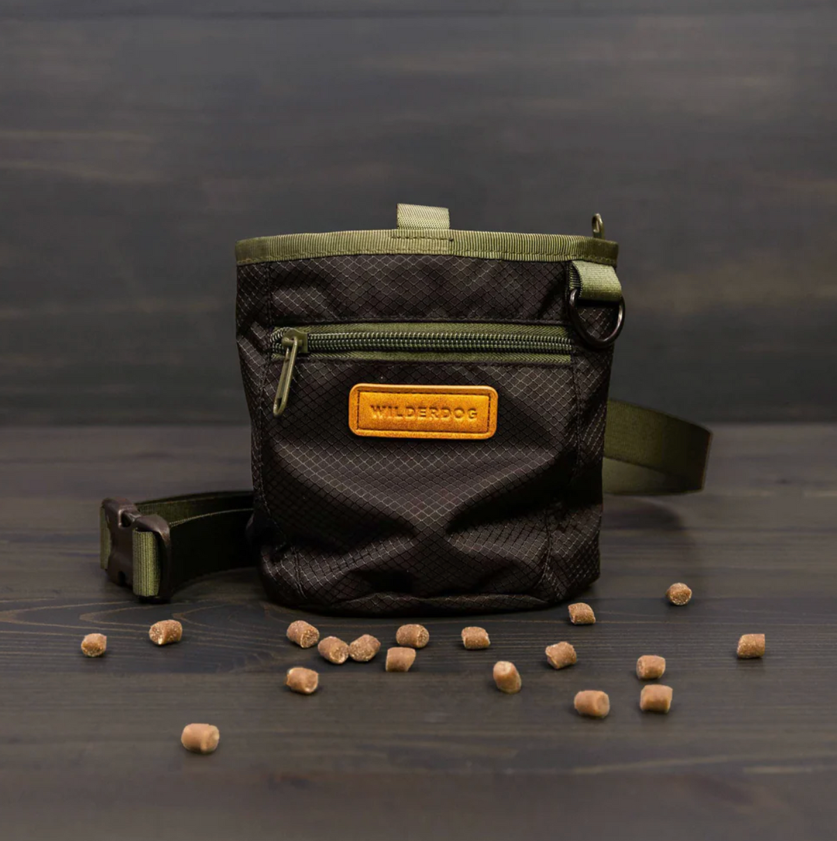 A black textured dog sling bag with olive green straps and a tan leather brand patch reading 'WILDERDOG'. The bag is displayed on a dark wooden surface with scattered dog treats in front of it.