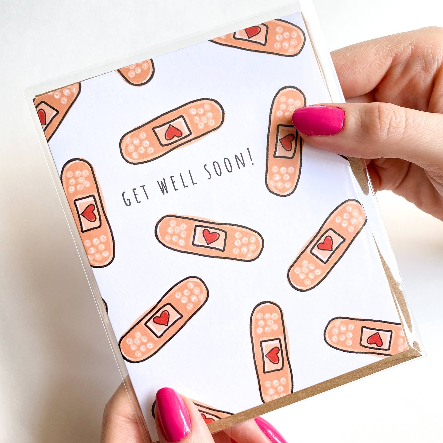 Card with bandage pattern and 'Get Well Soon!' text held by a hand with pink nail polish.
