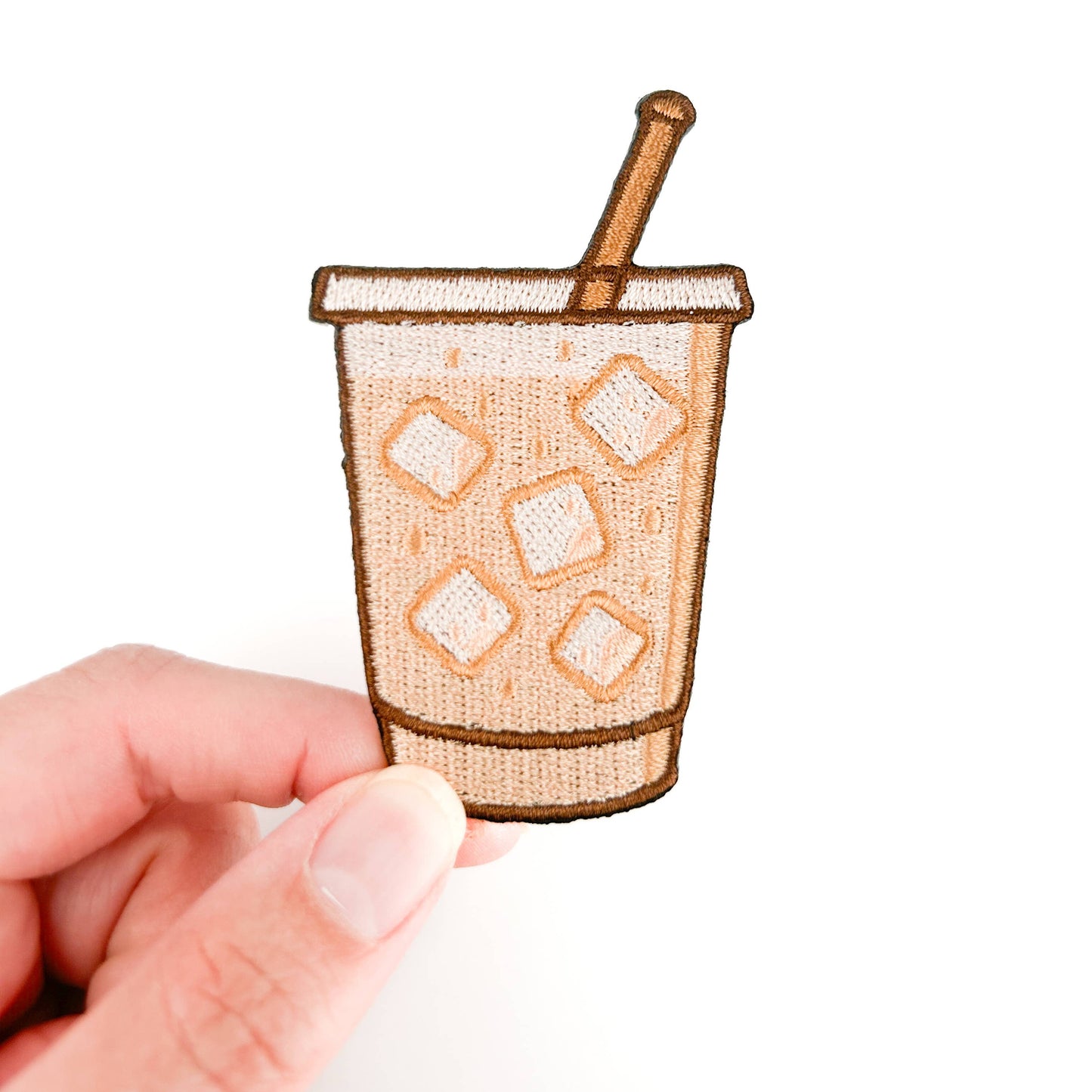 Small embroidered patch of a coffee cup with ice and a straw held by a hand on a white background