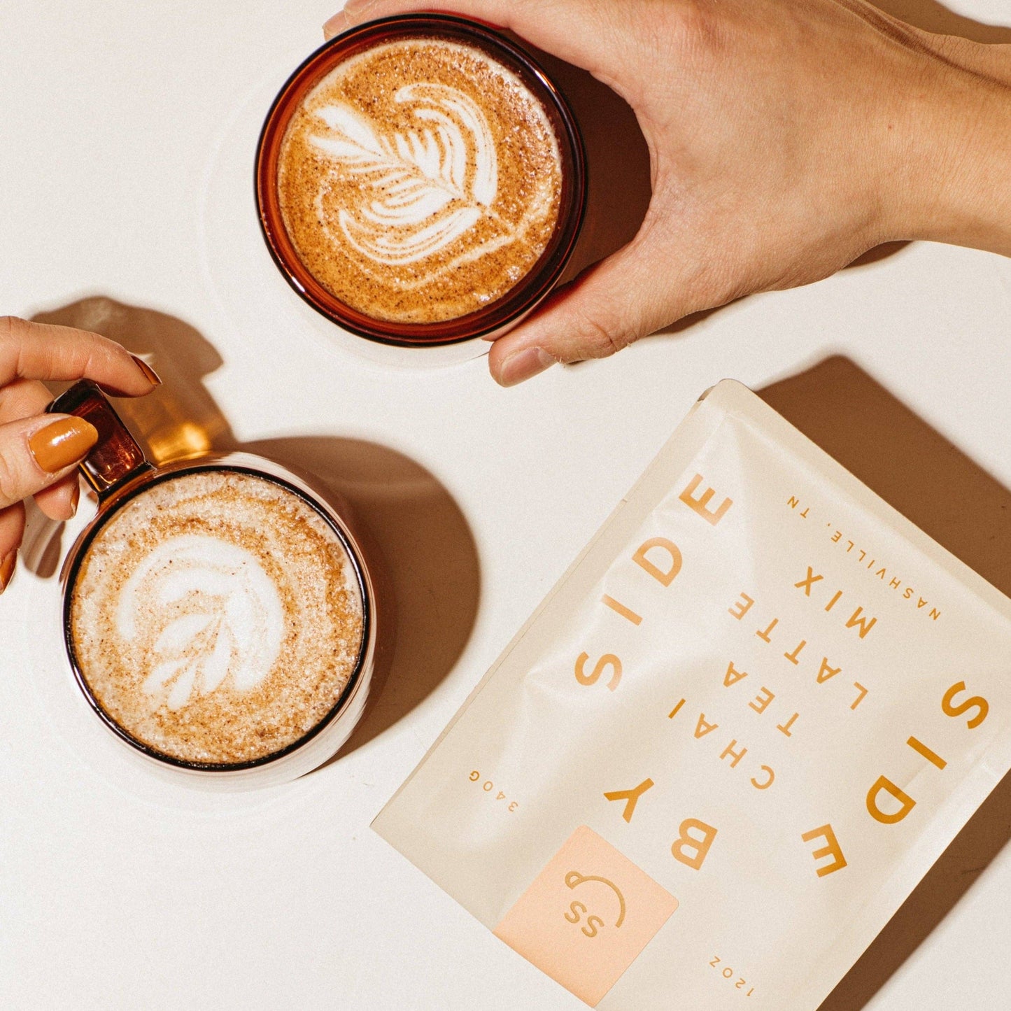 Two hands holding mugs of cappuccino with a Side by Side tea and coffee mix package on a light background.