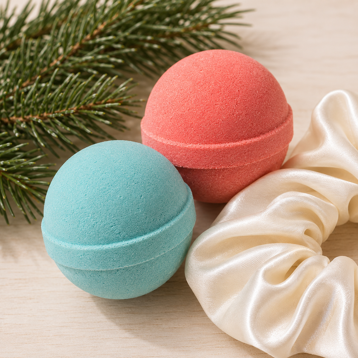 Two round bath bomb spheres, one light aqua blue and one coral pink, styled with green pine branches and a cream satin scrunchie on a light wood surface.