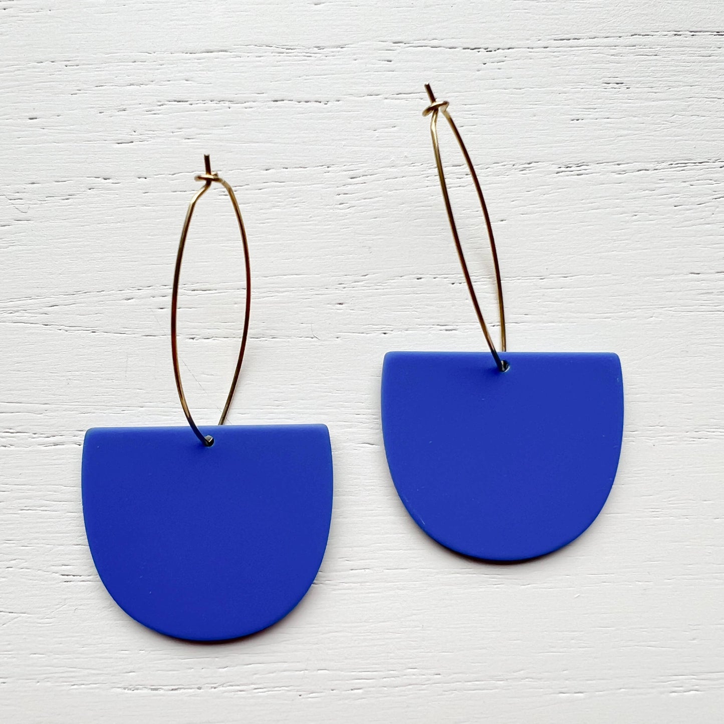 A pair of statement earrings featuring matte royal blue semi-circular drop pendants suspended from thin gold wire hoops, displayed on a white wood-textured background.
