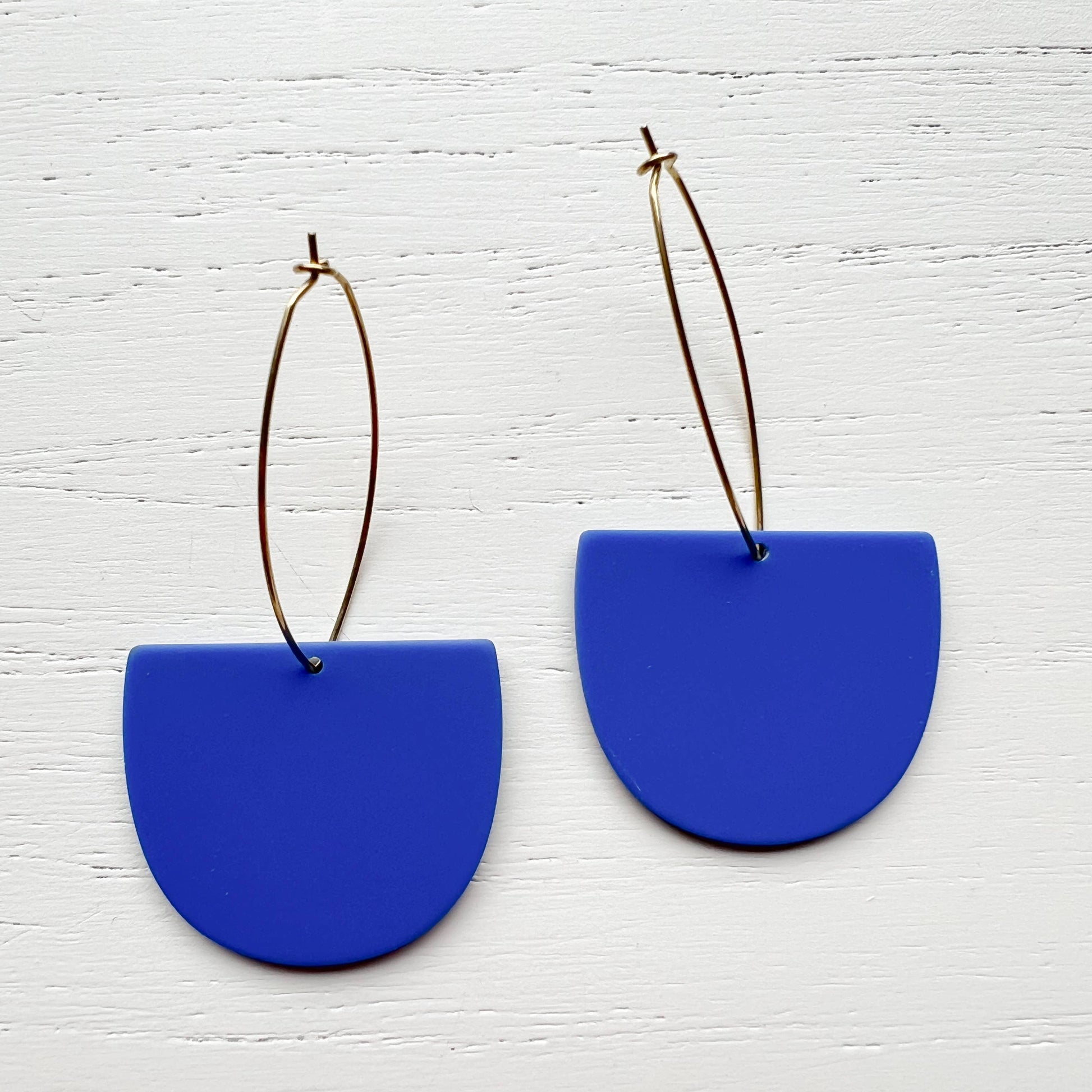 A pair of statement earrings featuring matte royal blue semi-circular drop pendants suspended from thin gold wire hoops, displayed on a white wood-textured background.
