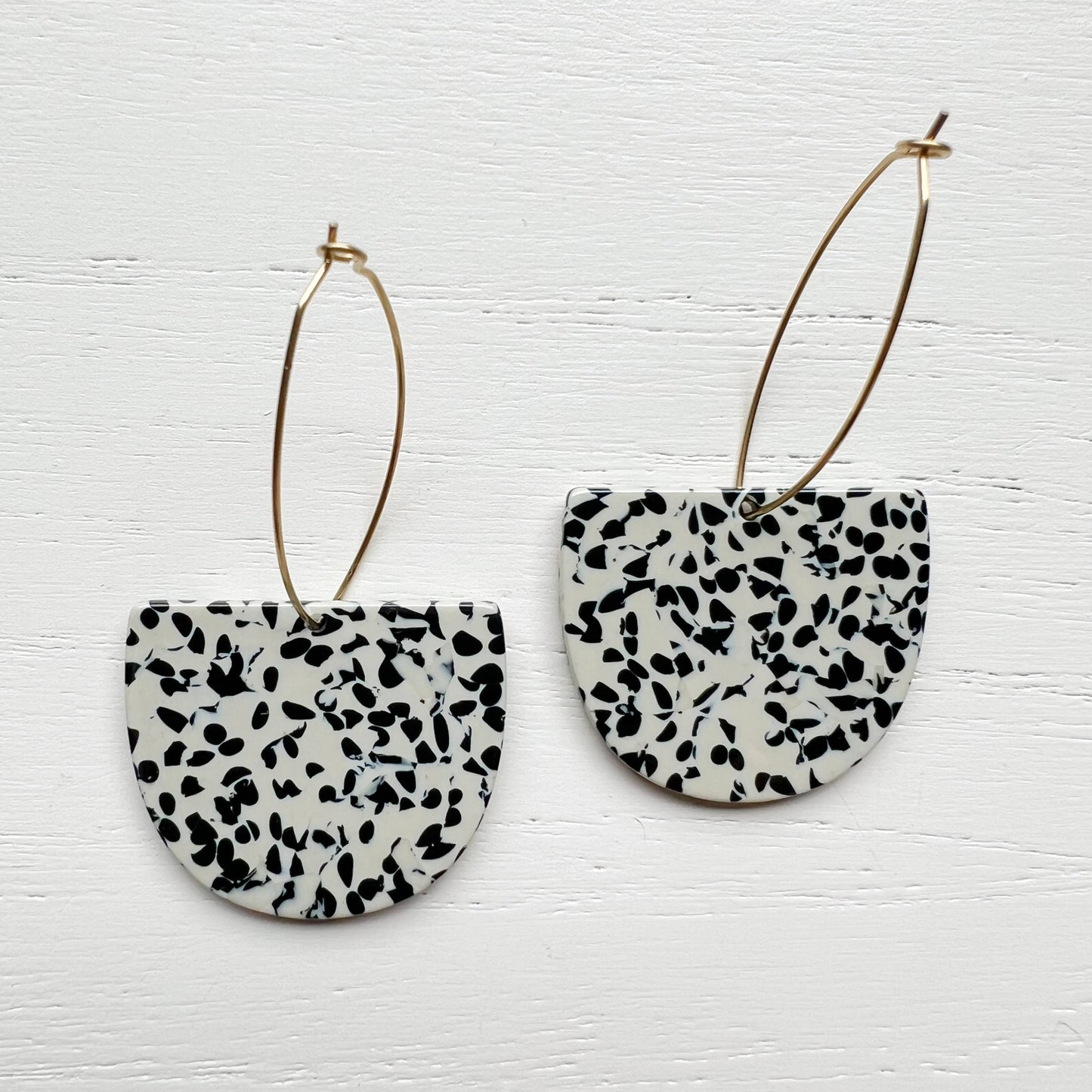 A pair of black and white speckled semi-circular drop earrings with thin gold hoop wires, displayed on a white painted wood background.