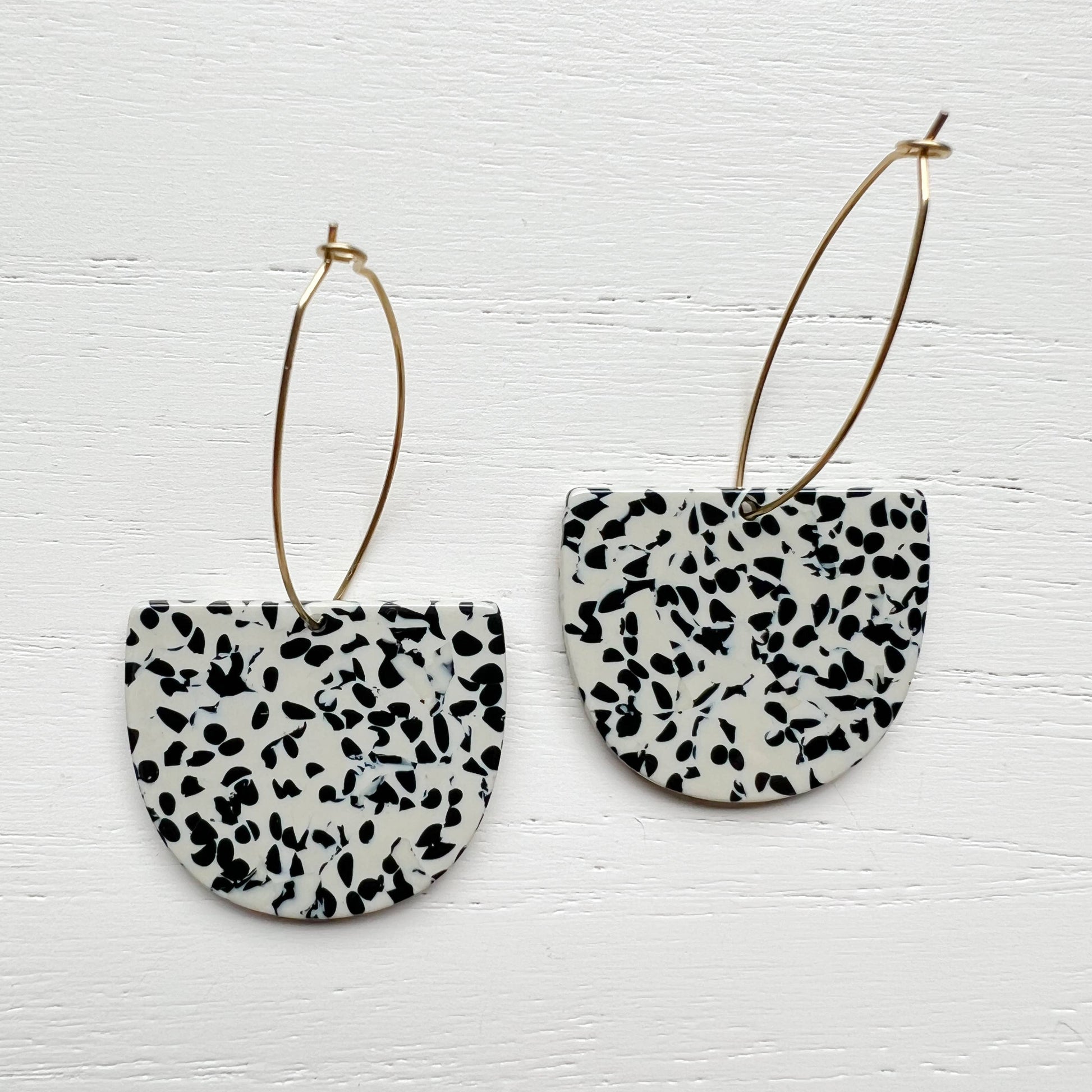 A pair of black and white speckled semi-circular drop earrings with thin gold hoop wires, displayed on a white painted wood background.