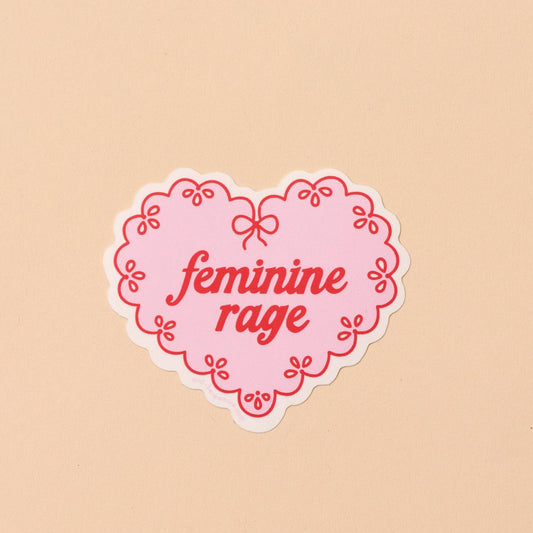 Heart-shaped sticker with 'feminine rage' text on a beige background