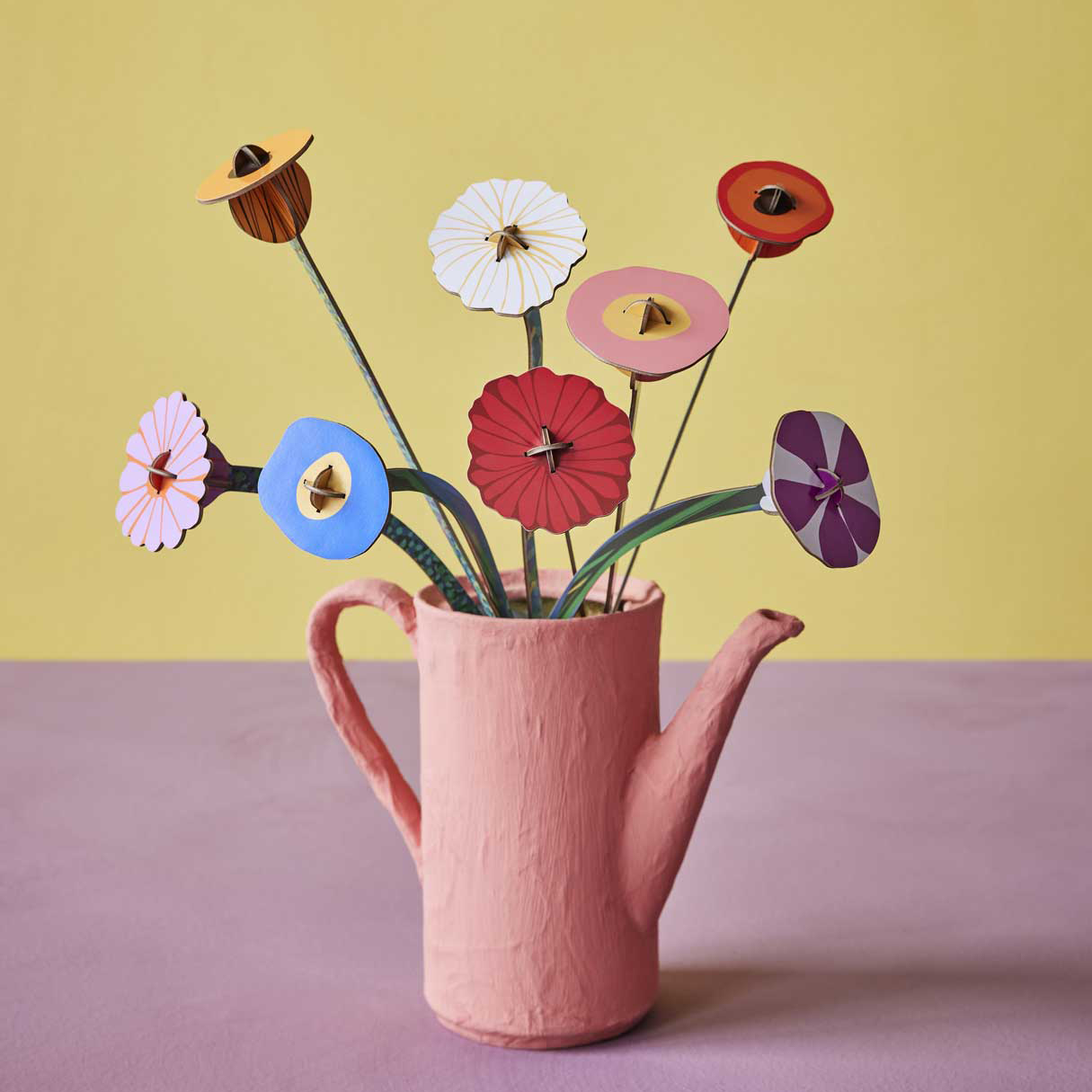Pink teapot with colorful paper flowers against a yellow background