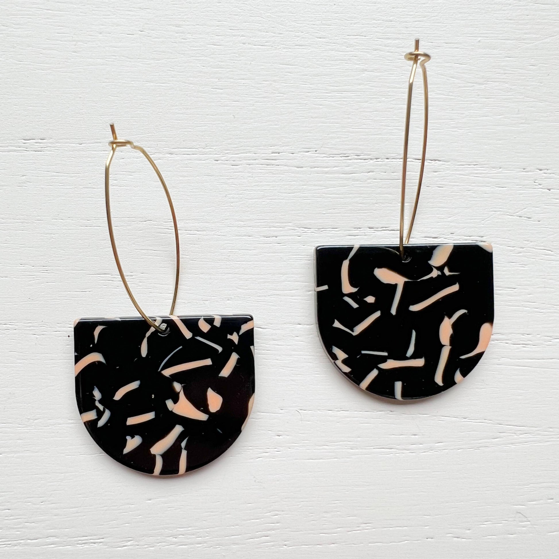 A pair of geometric drop earrings featuring black terrazzo-patterned semi-circular pendants with gold wire hoops, displayed against a white painted wood background.