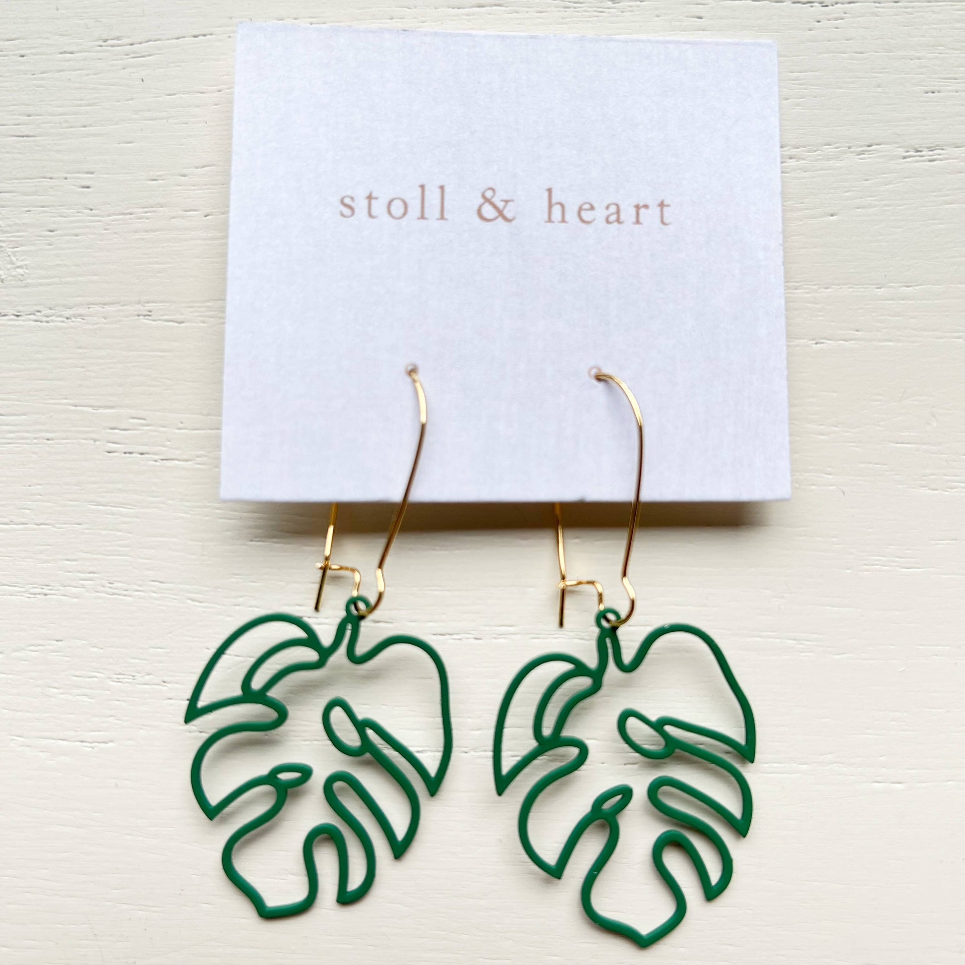 Green monstera leaf outline dangle earrings with gold hooks, displayed on a white card labeled 'stoll & heart' against a light wood background.