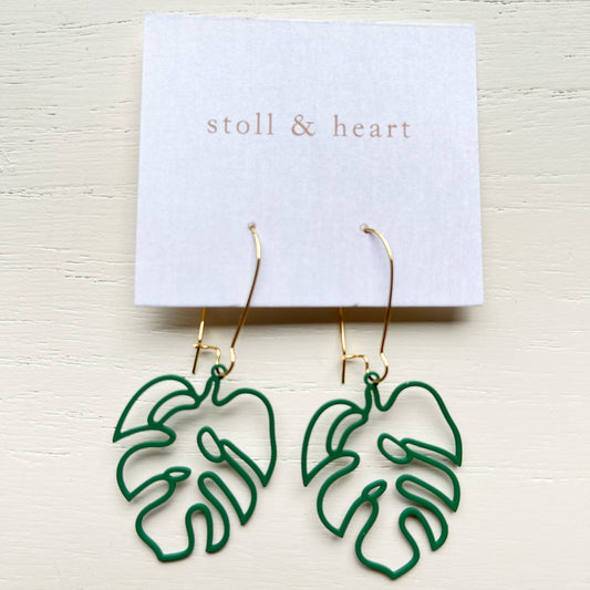Green monstera leaf outline dangle earrings with gold hooks, displayed on a white card labeled 'stoll & heart' against a light wood background.