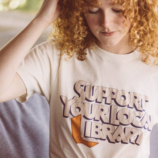 Person wearing a 'Support Your Local Library' t-shirt, sitting on a couch.