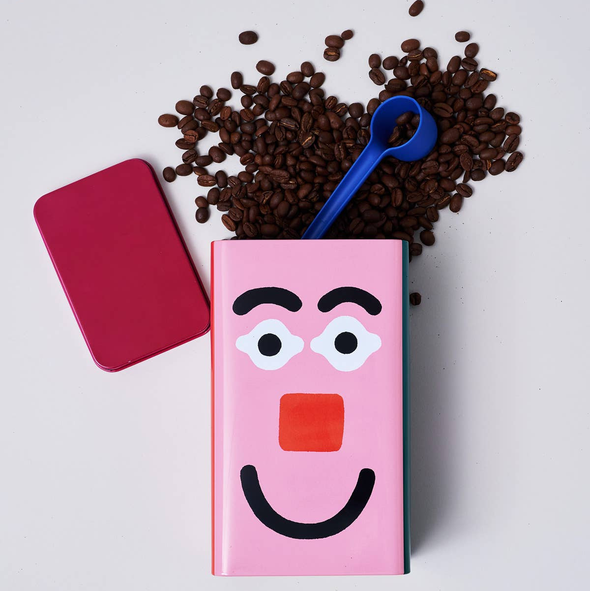 Pink container with cartoon face design, surrounded by coffee beans and a blue scoop on a light gray background.