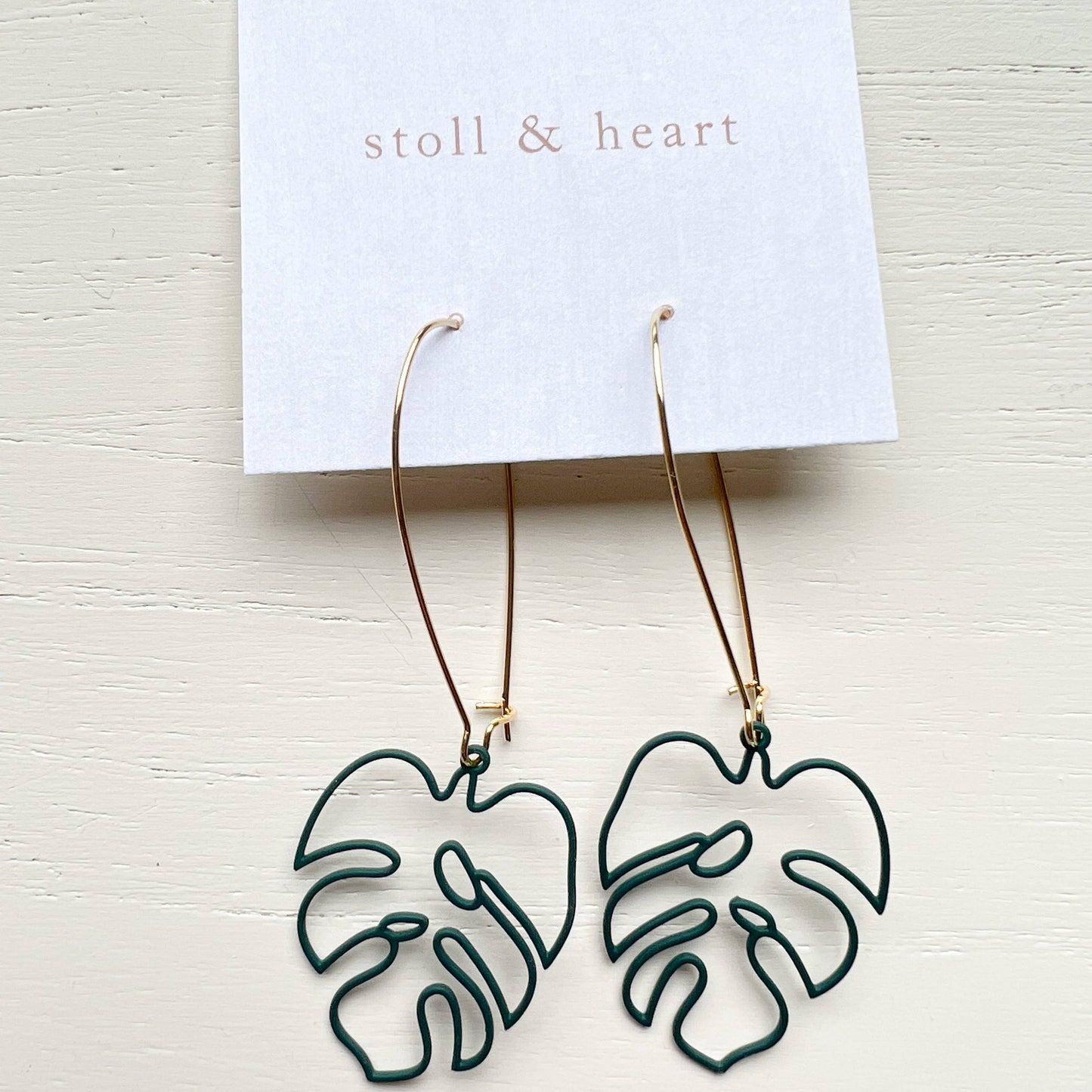 A pair of dangling hoop earrings with dark green monstera leaf cutouts and gold-tone wire hooks, displayed on a white card labeled 'stoll & heart' against a light wood background.