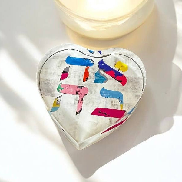 Heart-shaped decorative item with colorful letters next to a candle on a white surface