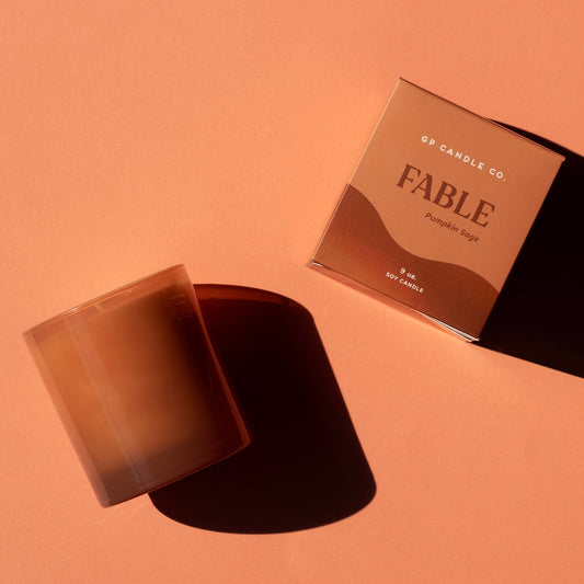 A 5-oz soy candle by FP Candle Co. in a translucent amber glass jar, shown next to its matching square packaging box on a warm peach background.
