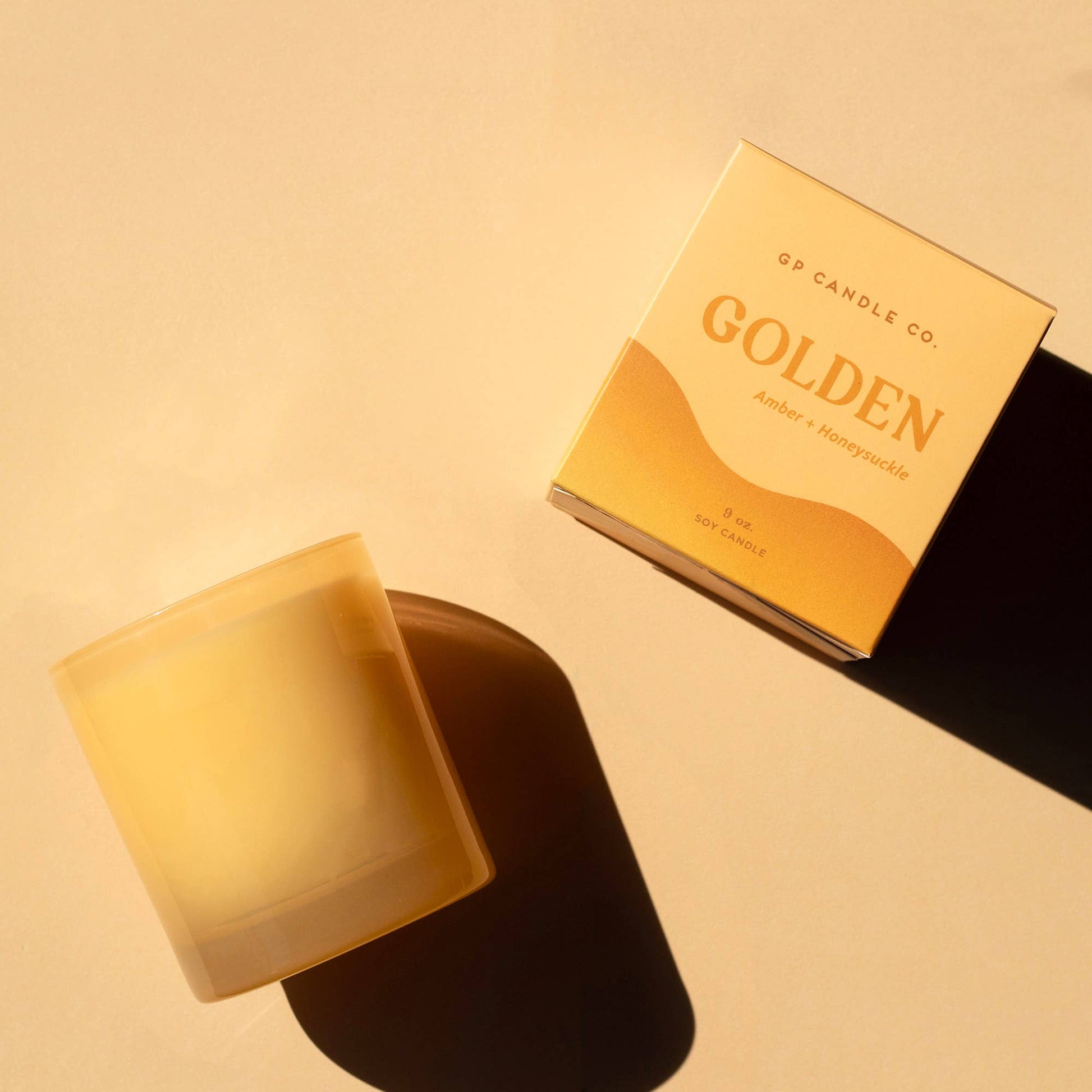 A candle in a frosted glass jar sits beside its branded box on a warm beige surface. The packaging reads 'GP CANDLE CO.