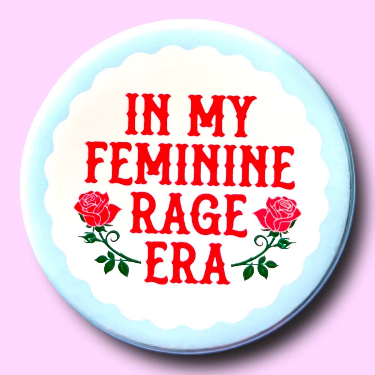 Button badge with 'In my feminine rage era' text and roses on a light blue border against a pink background