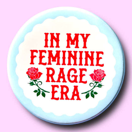 Button badge with 'In my feminine rage era' text and roses on a light blue border against a pink background