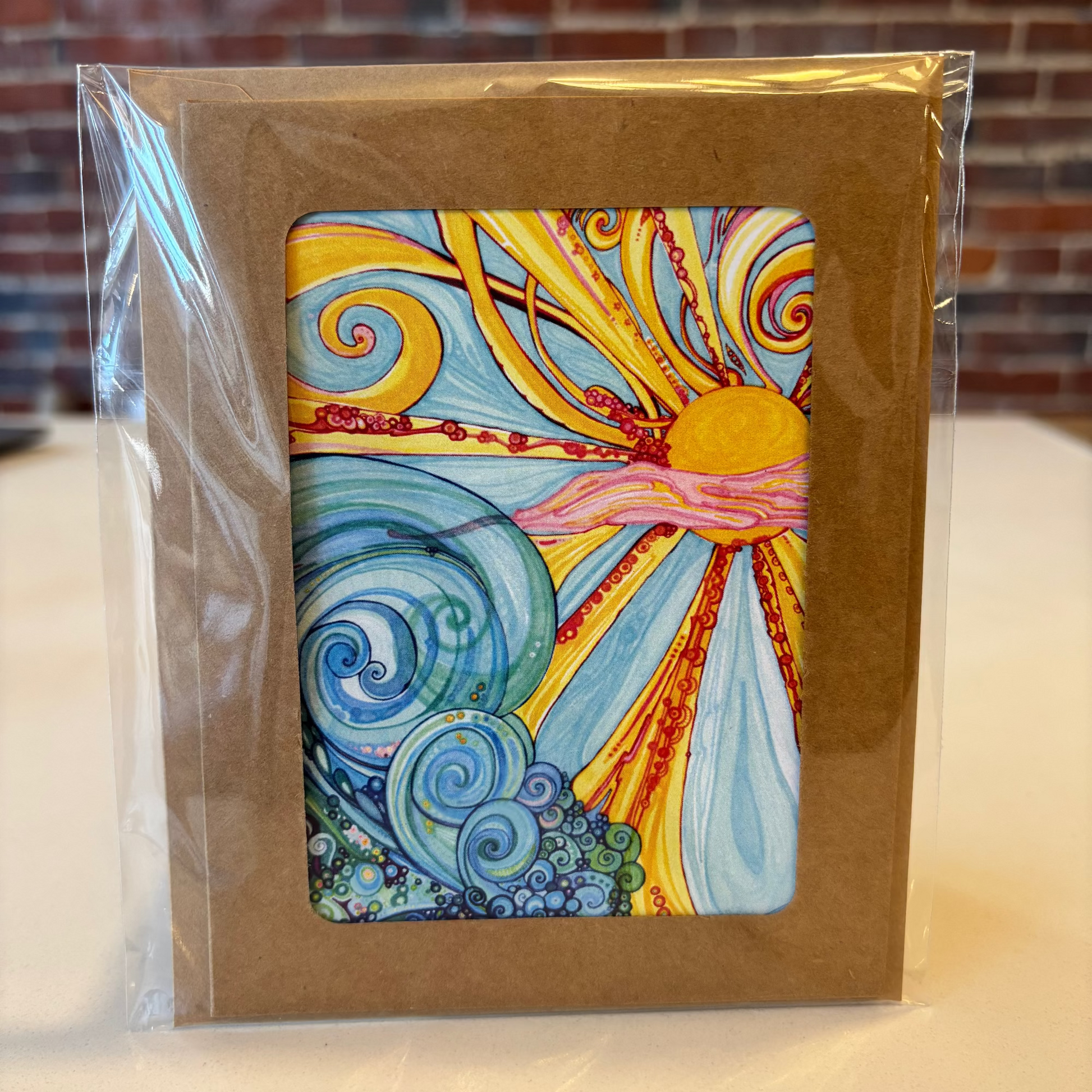 Colorful abstract design on a card in clear packaging with a brown border, against a brick wall background.