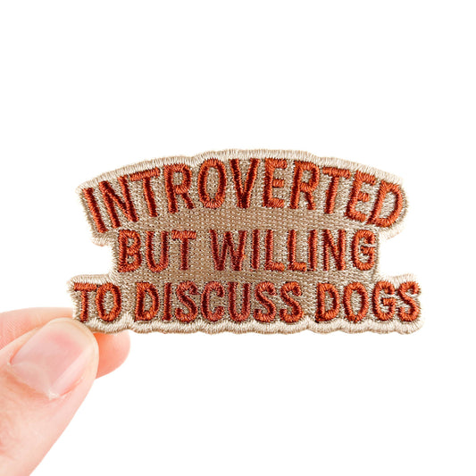 Patch with text 'Introverted but willing to discuss dogs' held by a hand on a white background
