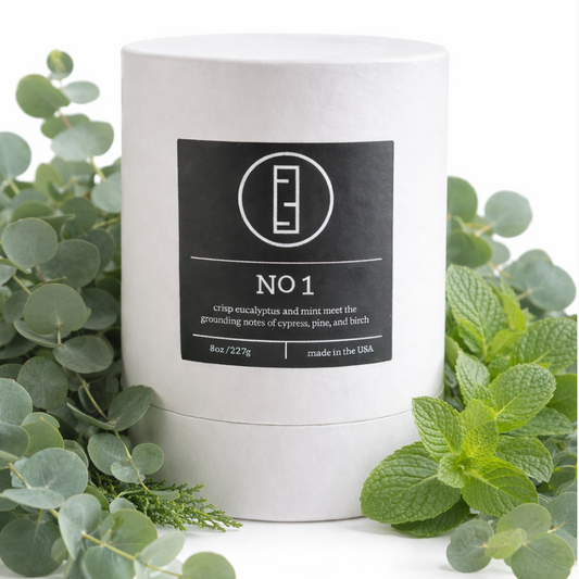 White candle with a black label surrounded by green leaves on a white background