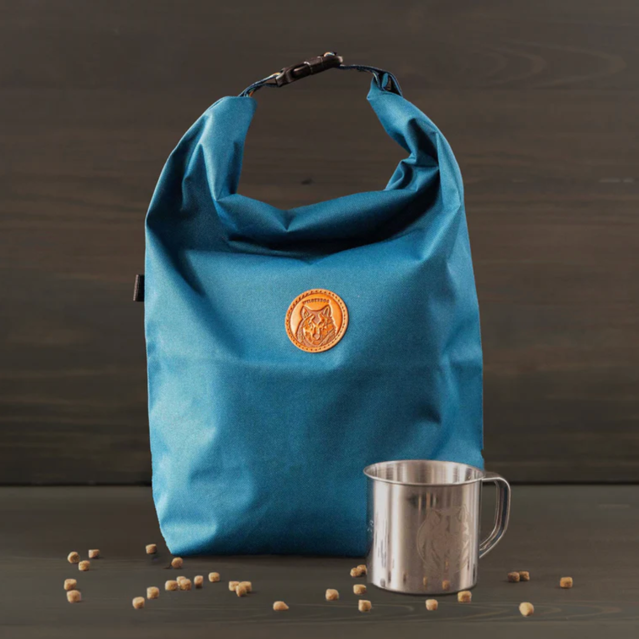 A blue waterproof bag with a circular tan logo patch on the front, standing on a dark wooden surface next to a stainless steel mug with scattered coffee beans in front.