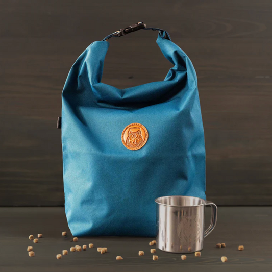 A blue waterproof bag with a circular tan logo patch on the front, standing on a dark wooden surface next to a stainless steel mug with scattered coffee beans in front.
