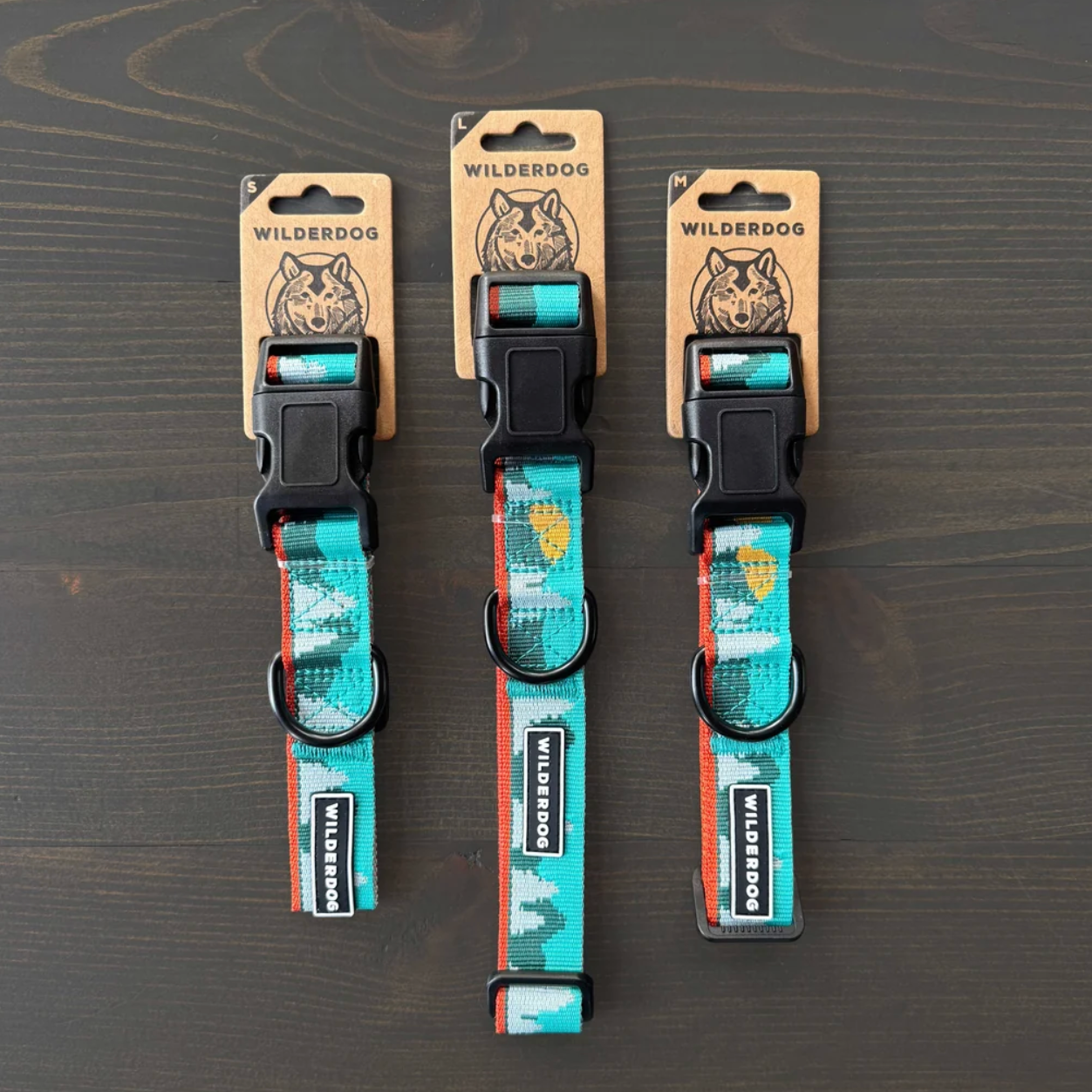 Three dog collars with black plastic quick-release buckles and metal rings, hanging from kraft paper Wilderdog brand tags on a dark wood surface.