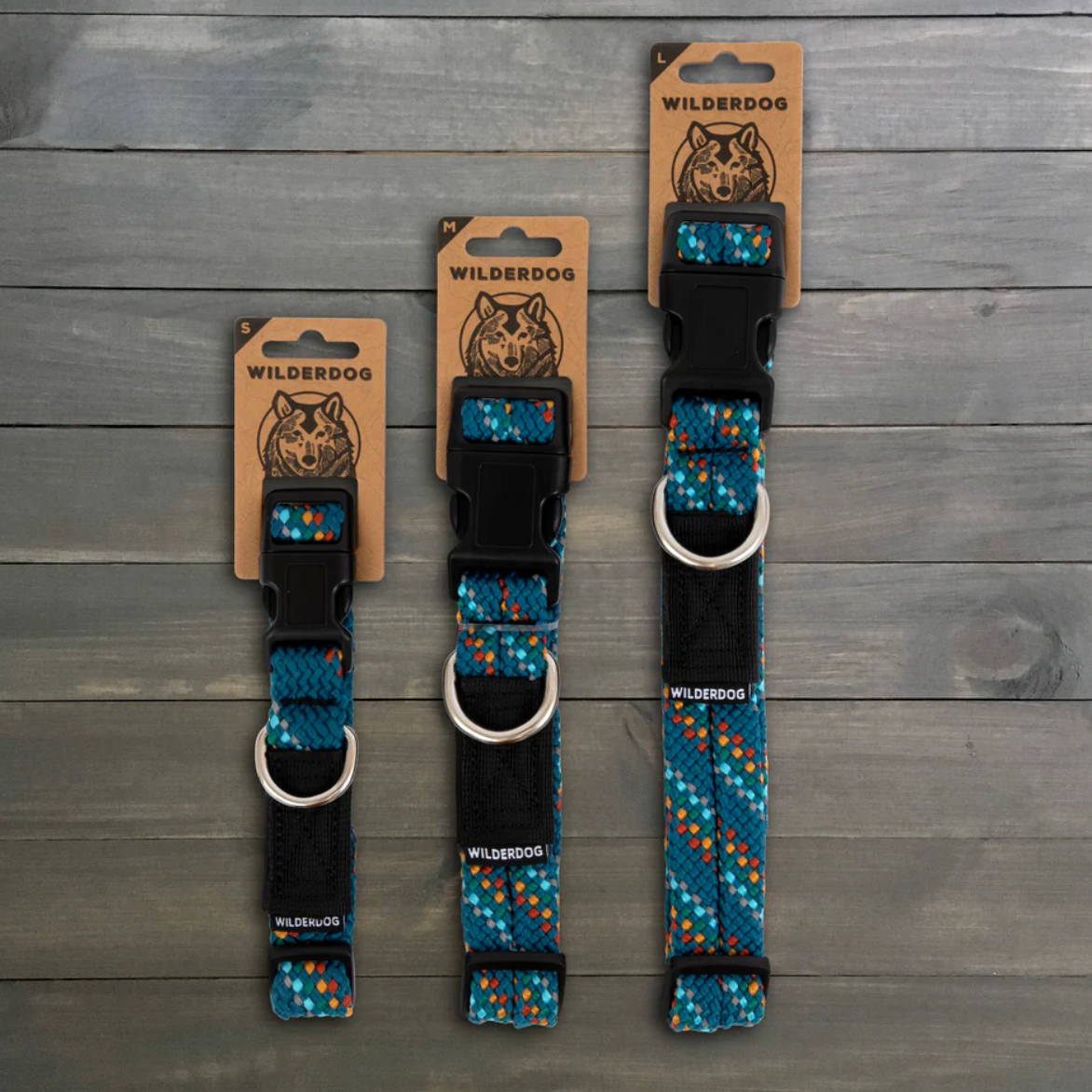 Three WildDog pet collars in different sizes displayed vertically on a gray wood plank background. Each collar has a blue, black, and orange patterned webbing strap, a black plastic buckle, and a silver metal D-ring.