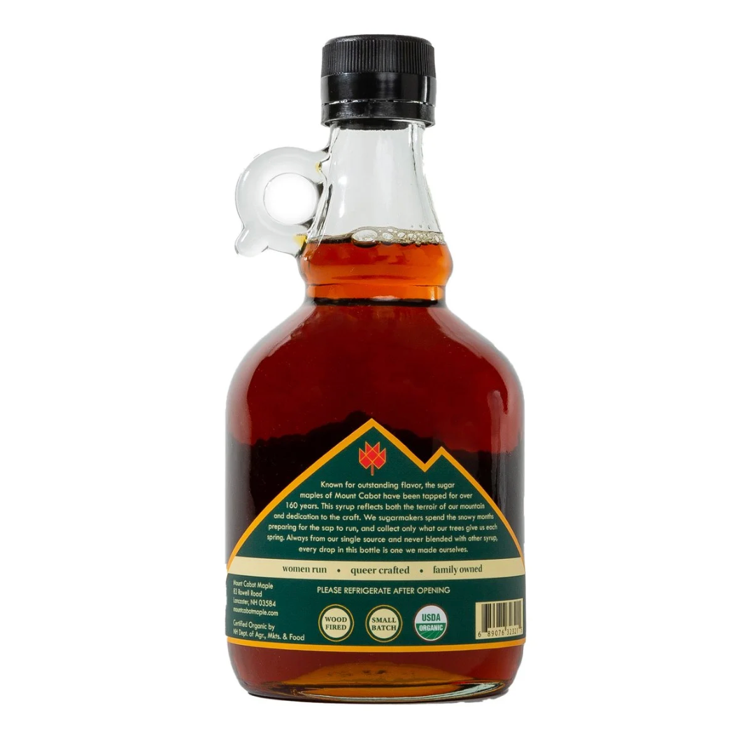 A bottle of maple syrup with a clear glass body, amber liquid, and a black cap, shown from the back with a green and gold label on a white background.