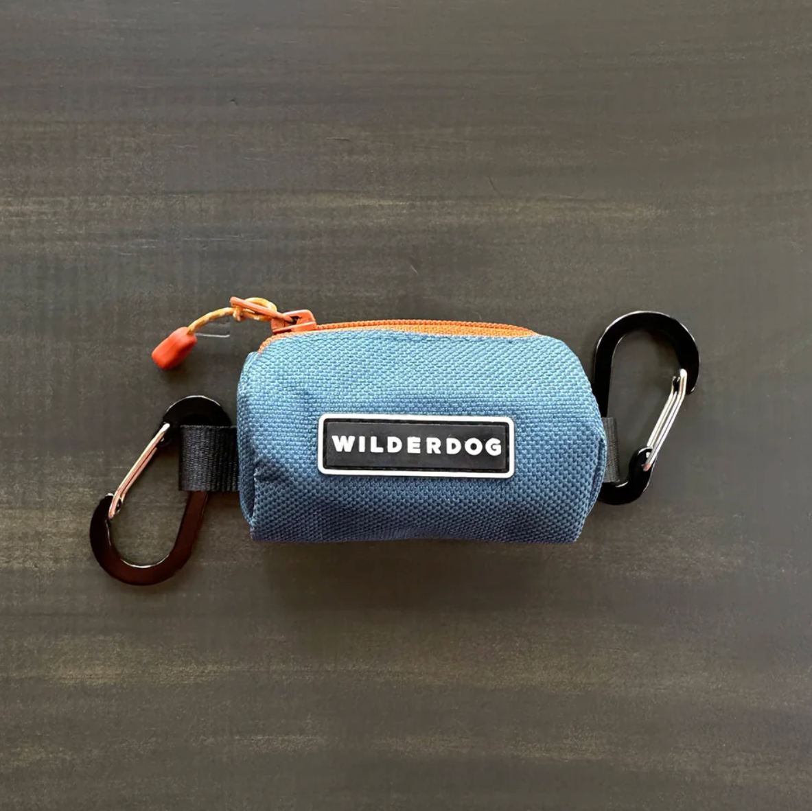 A blue canvas zippered pouch with a black and white 'WILDERDOG' brand patch on the front, shown on a dark gray textured background. The pouch is equipped with a black carabiner on one side, a brown strap on the other, and an orange zipper pull.