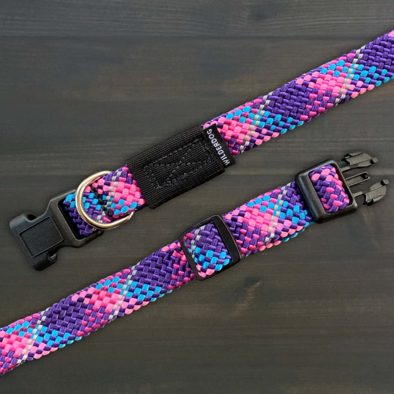 A woven pet collar with a vibrant pink, purple, and blue pattern, featuring black plastic buckles and a black D-ring, displayed on a dark wood background.