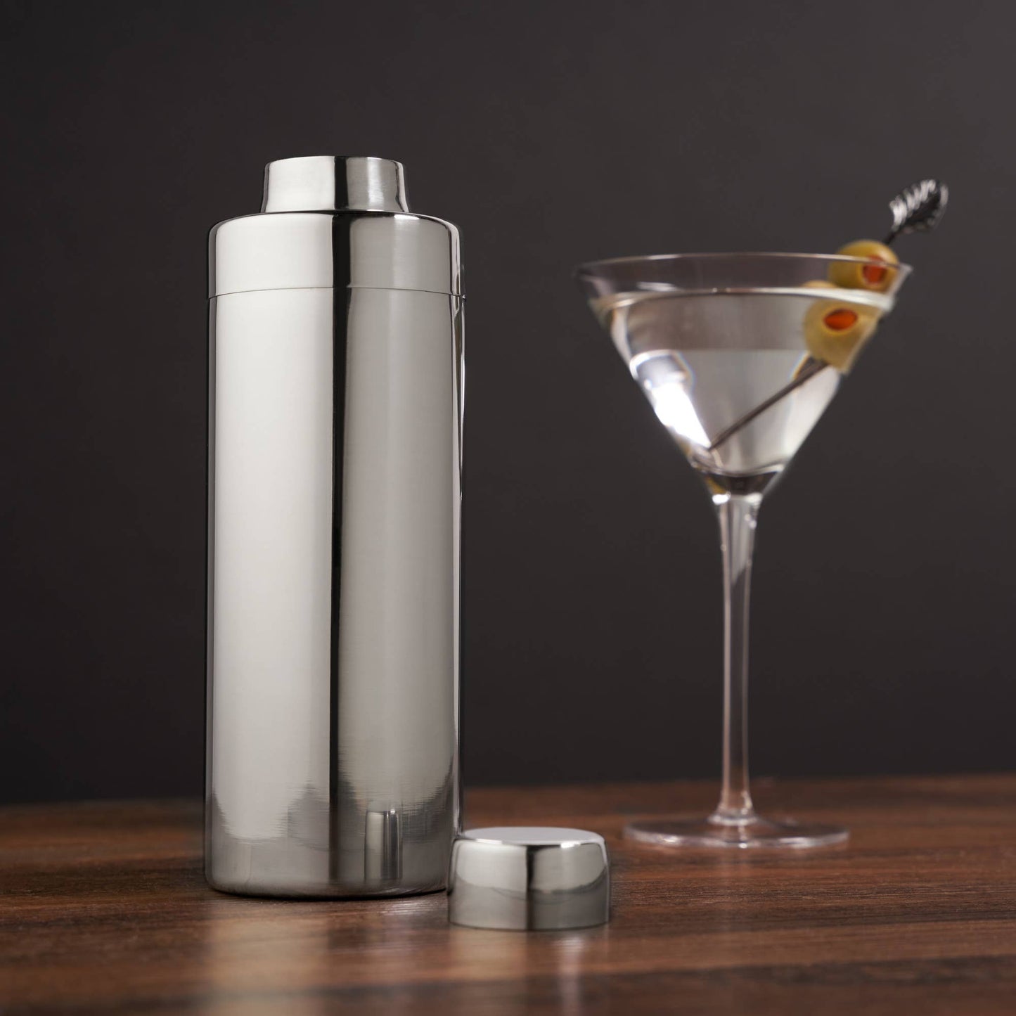 Metal cocktail shaker next to a martini glass with olives on a wooden surface.
