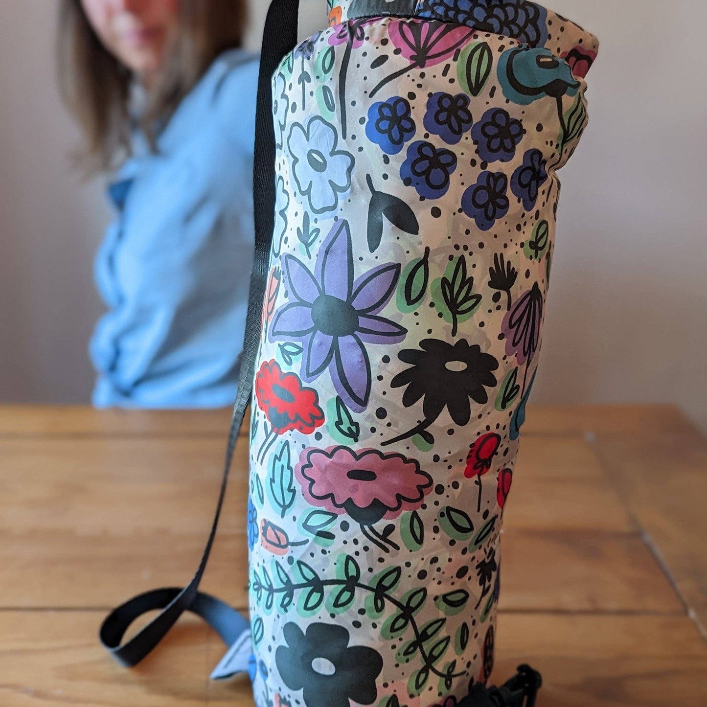 Floral-patterned water bottle holder on a wooden surface with a blurred person in the background