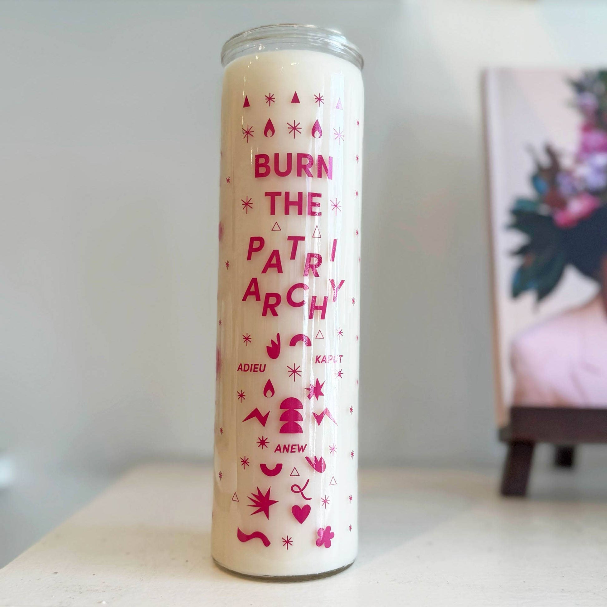 A tall glass jar candle with a cream-colored wax body and bright pink printed text and symbols reading 'BURN THE PATRIARCHY' and 'ADIEU KAPOT ANEW',
