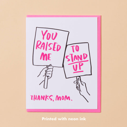 Card with hands holding signs saying 'You raised me to stand up' and 'Thanks, Mom.' on a beige background.