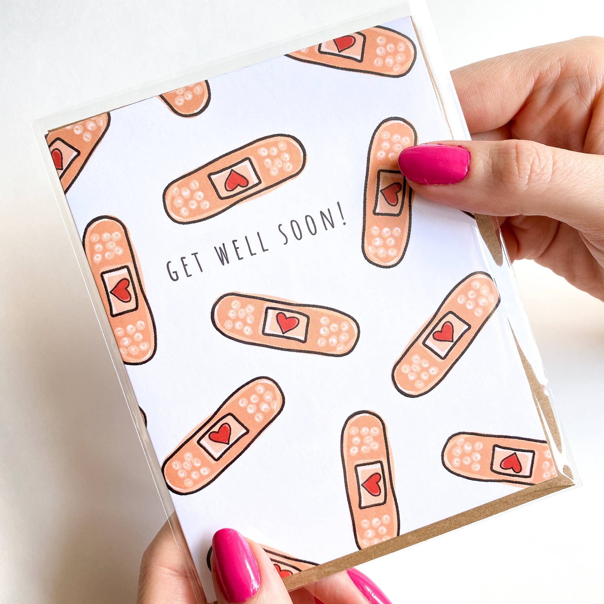 Card with bandage pattern and 'Get Well Soon!' text held by a hand with pink nail polish.