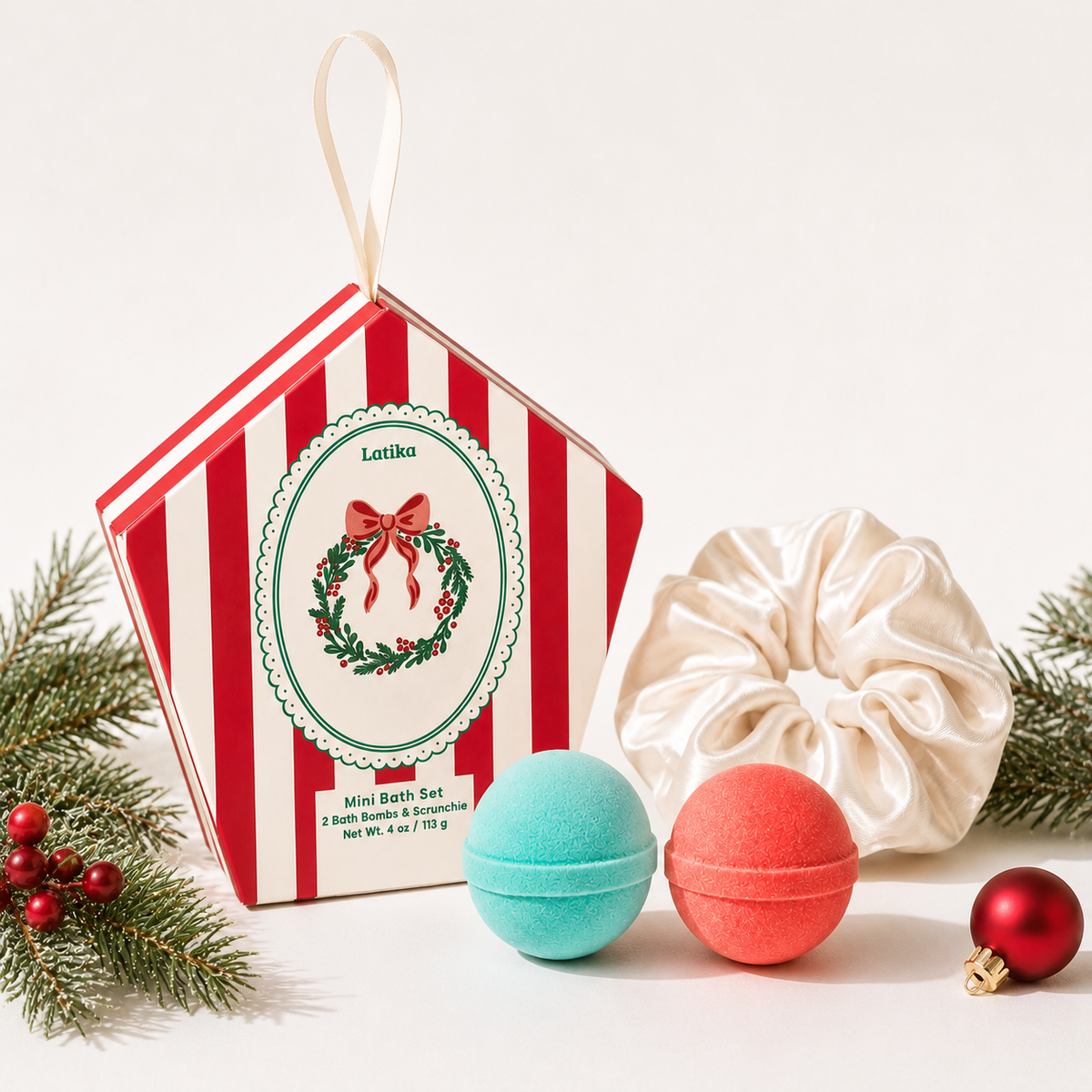 A Christmas-themed bath set gift box by Lotika, shown with a cream satin washcloth and two bath bombs in mint green and coral red, styled with pine branches and a red ornament on a white background.