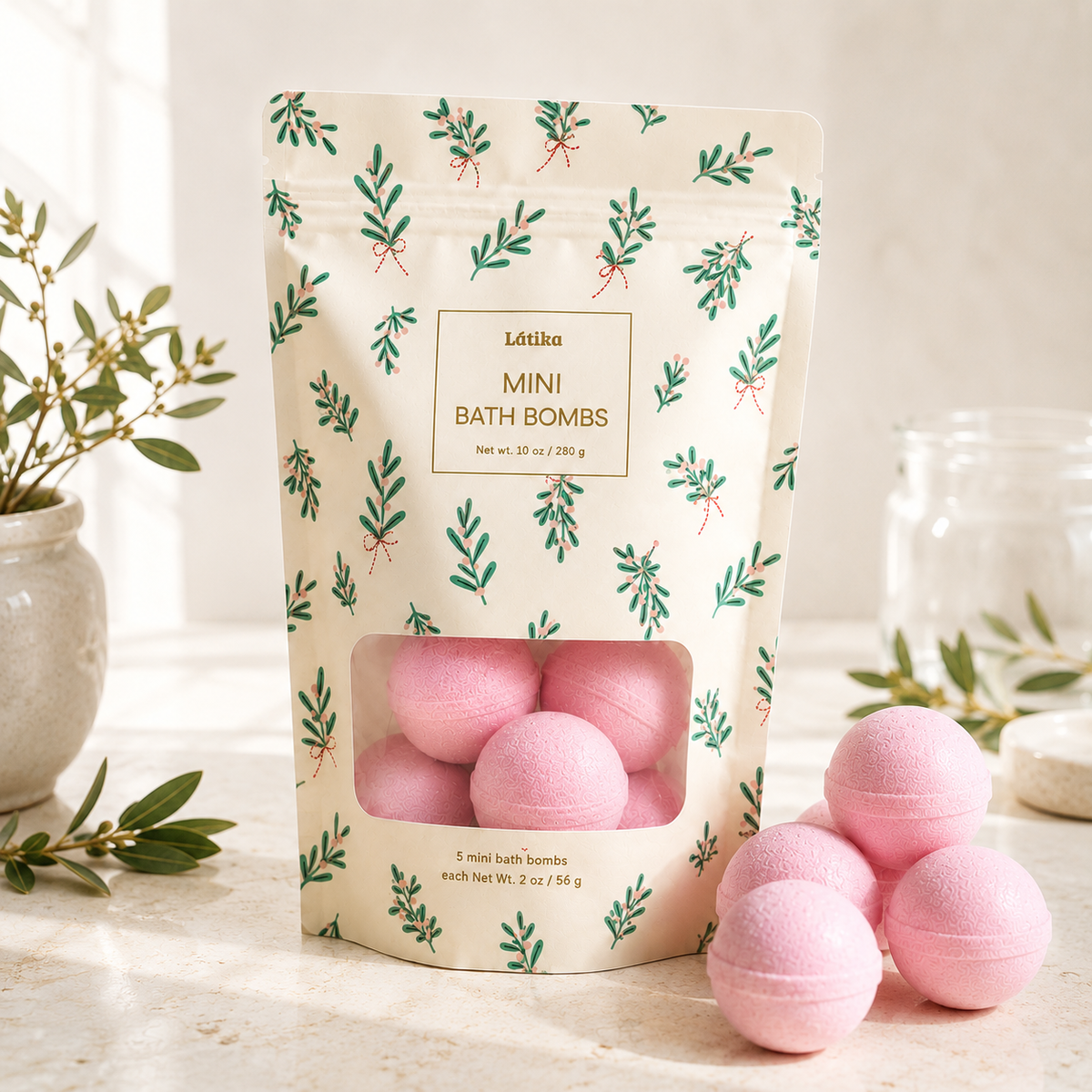 A stand-up pouch of Loteria Mini Bath Bombs with a clear window showing pink bath bombs inside, set on a light marble surface with several pink bath bombs beside it and soft decorative props in the background.