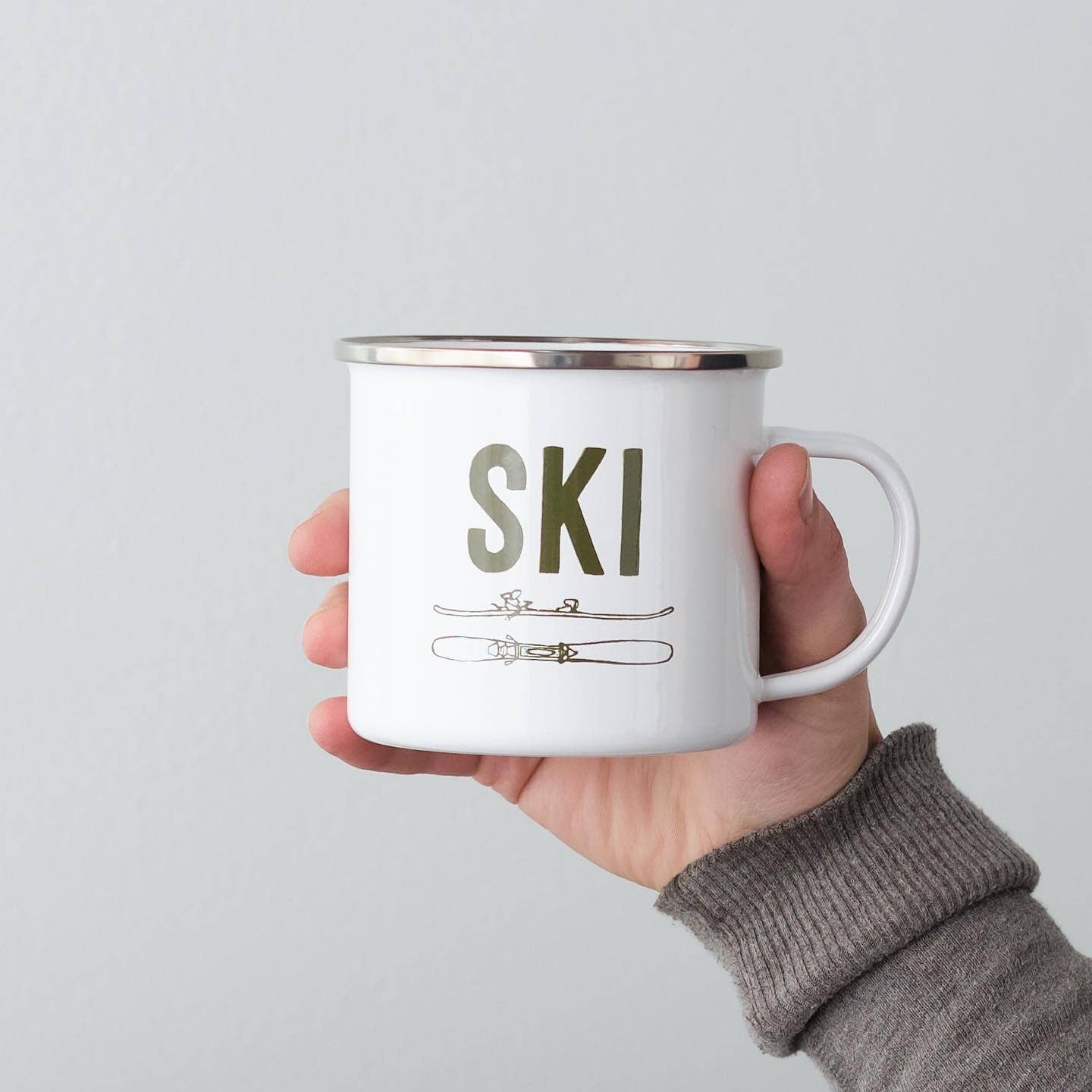 Hand holding a white mug with 'SKI' and ski illustrations on a light gray background