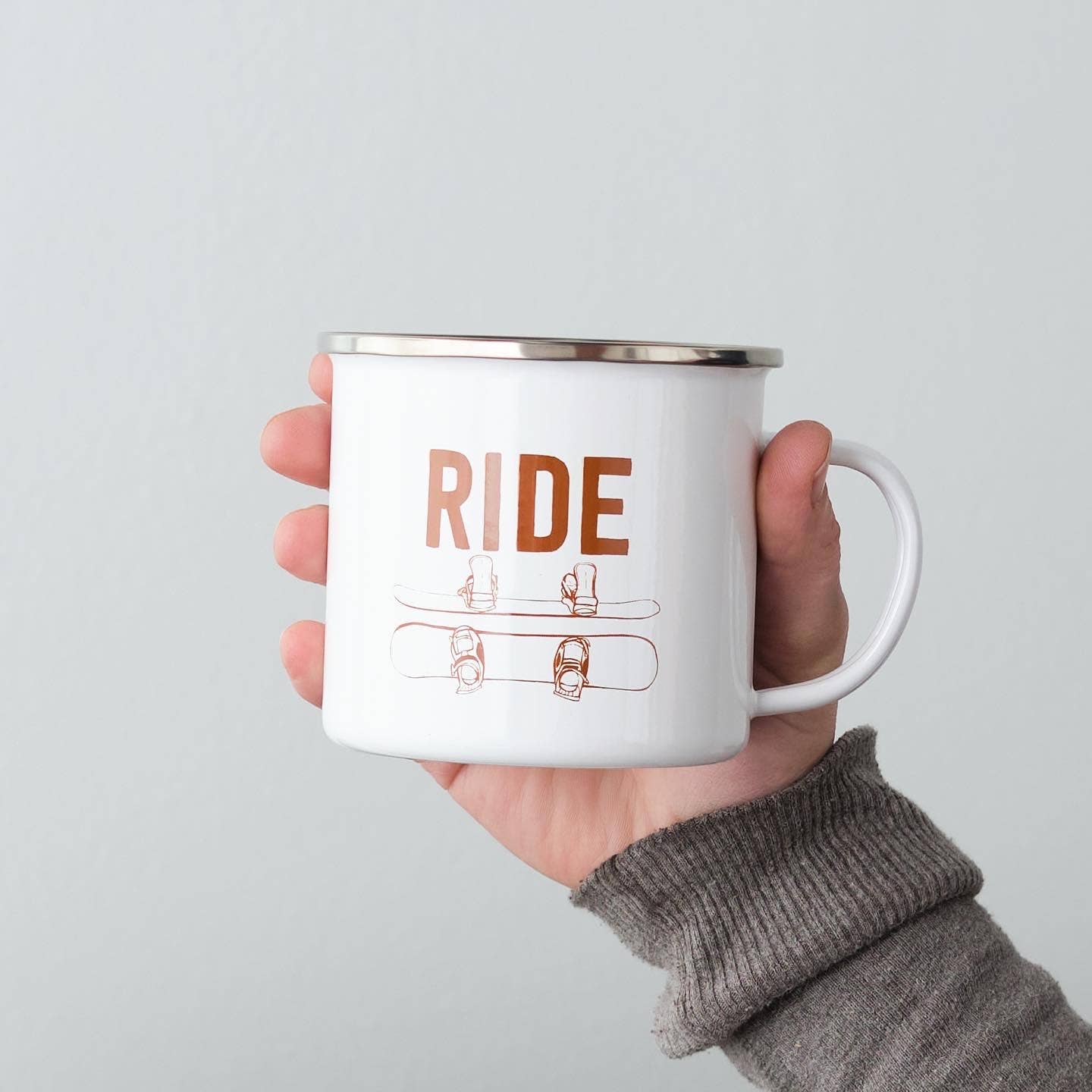 Hand holding a white mug with 'RIDE' and snowboarding graphics on a light gray background