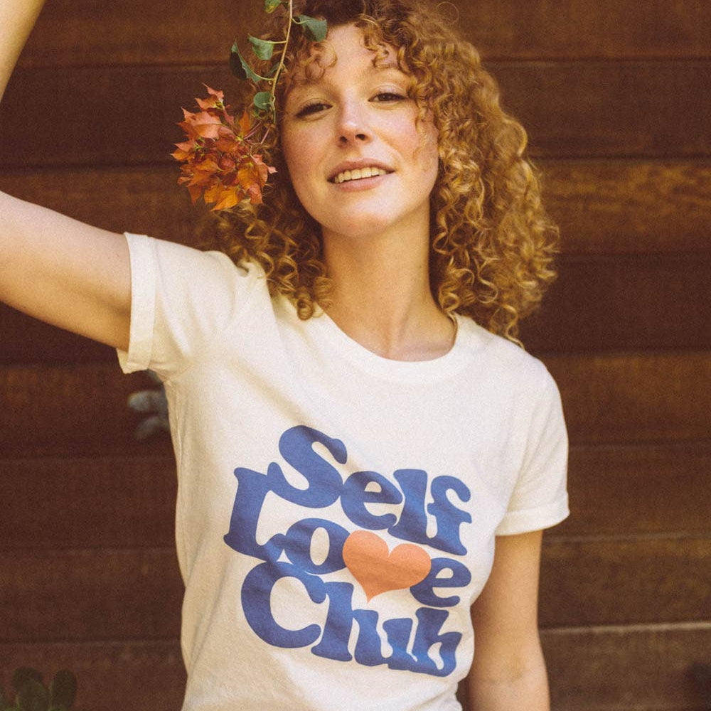 Woman wearing a 'Self Love Club' t-shirt with autumn leaves against a wooden background