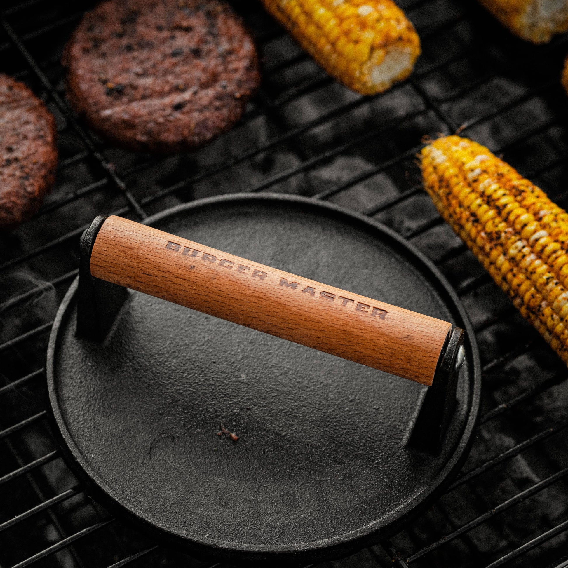 Burger press with wooden handle on a grill with burgers and corn