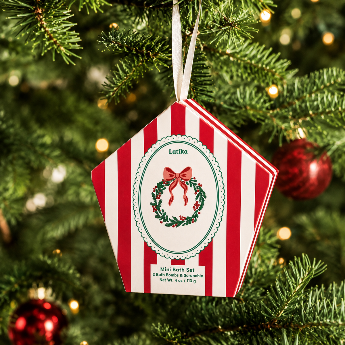 A red and white striped Christmas ornament package hanging from a green Christmas tree with warm bokeh lights. The package is hexagonal and features a wreath design in the center with the name 'Latika' at the top.