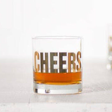 Glass with 'CHEERS' text filled with a brown liquid, blurred bottle in the background.